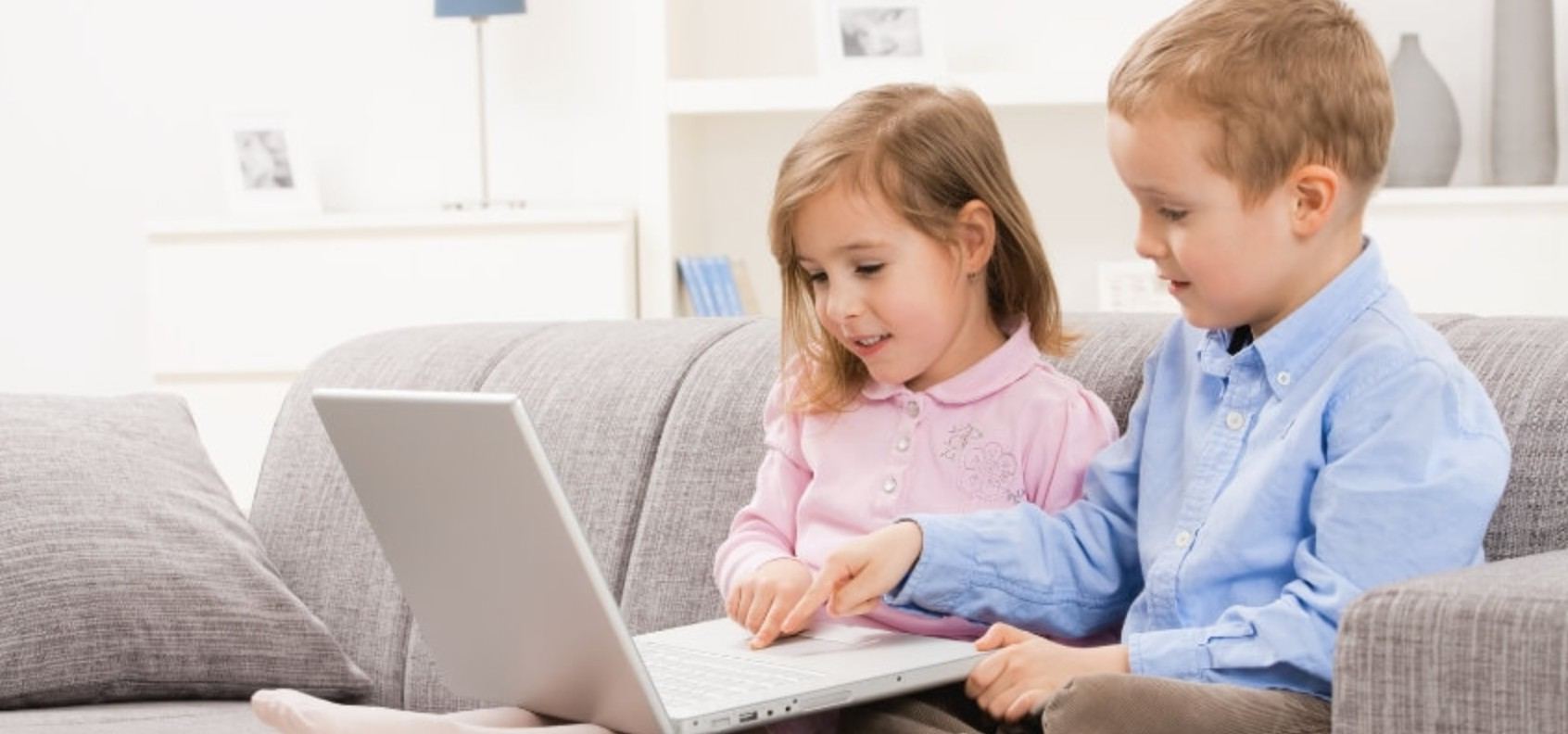 Двоє дошкільнят сидять на дивані та разом користуються ноутбуком під наглядом дорослих у домашньому середовищі