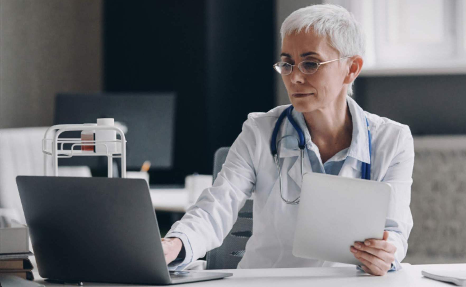 Лікарка працює за ноутбуком у медичному кабінеті та тримає планшет із даними пацієнта