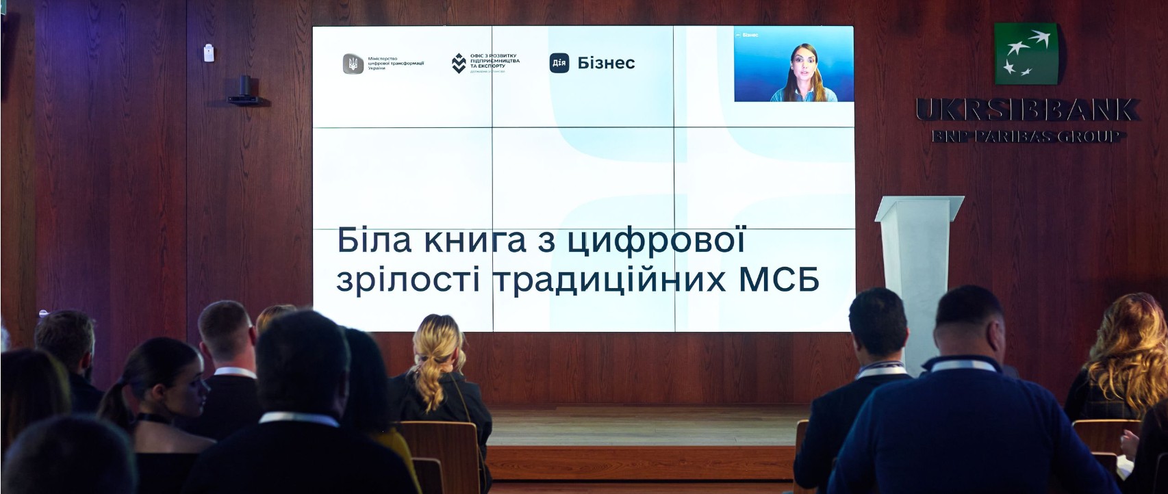 Зал з учасниками, перед якими на великому екрані демонструють презентацію «Біла книга з цифрової зрілості традиційних МСБ». На екрані також логотипи Мінцифри, Офісу з розвитку підприємництва та експорту, Дія.Бізнес і виступ спікерки онлайн.