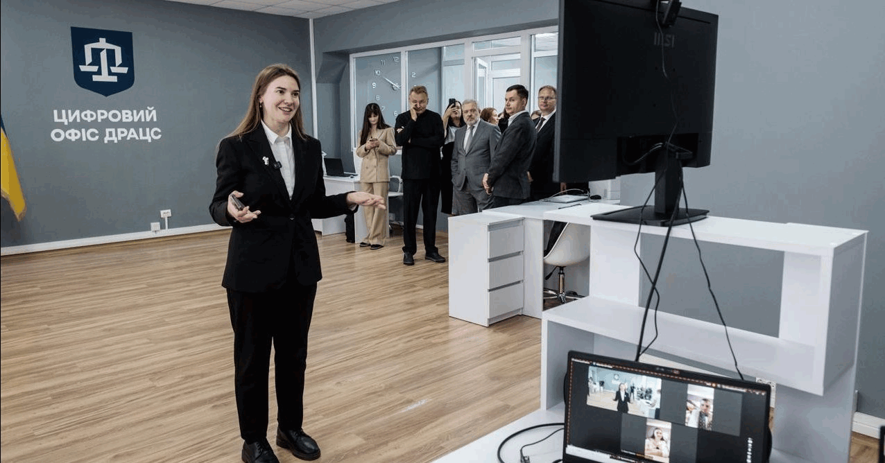 Сучасний офіс із написом «Цифровий офіс ДРАЦС» на стіні. На передньому плані — жінка у діловому костюмі проводить онлайн-презентацію або відеоконференцію, спілкуючись через великий монітор. На задньому плані група людей у діловому одязі спостерігає за подією. Інтер’єр світлий, сучасний, із дерев’яною підлогою та мінімалістичними меблями.