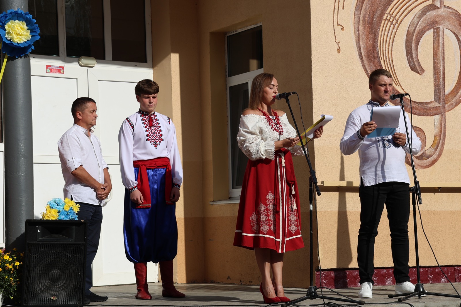 Урочистий мітинг біля будівлі закладу культури з барельєфом скрипкового ключа на фасаді. Біля мікрофонів стоять ведучі заходу: жінка в червоно-білому строї з червоним намистом і чоловік у вишиванці з папкою для виступів. Ліворуч на сцені стоять двоє чоловіків, один із яких одягнений в автентичний український костюм із вишиванкою, червоним поясом, синіми шароварами та чобітьми.