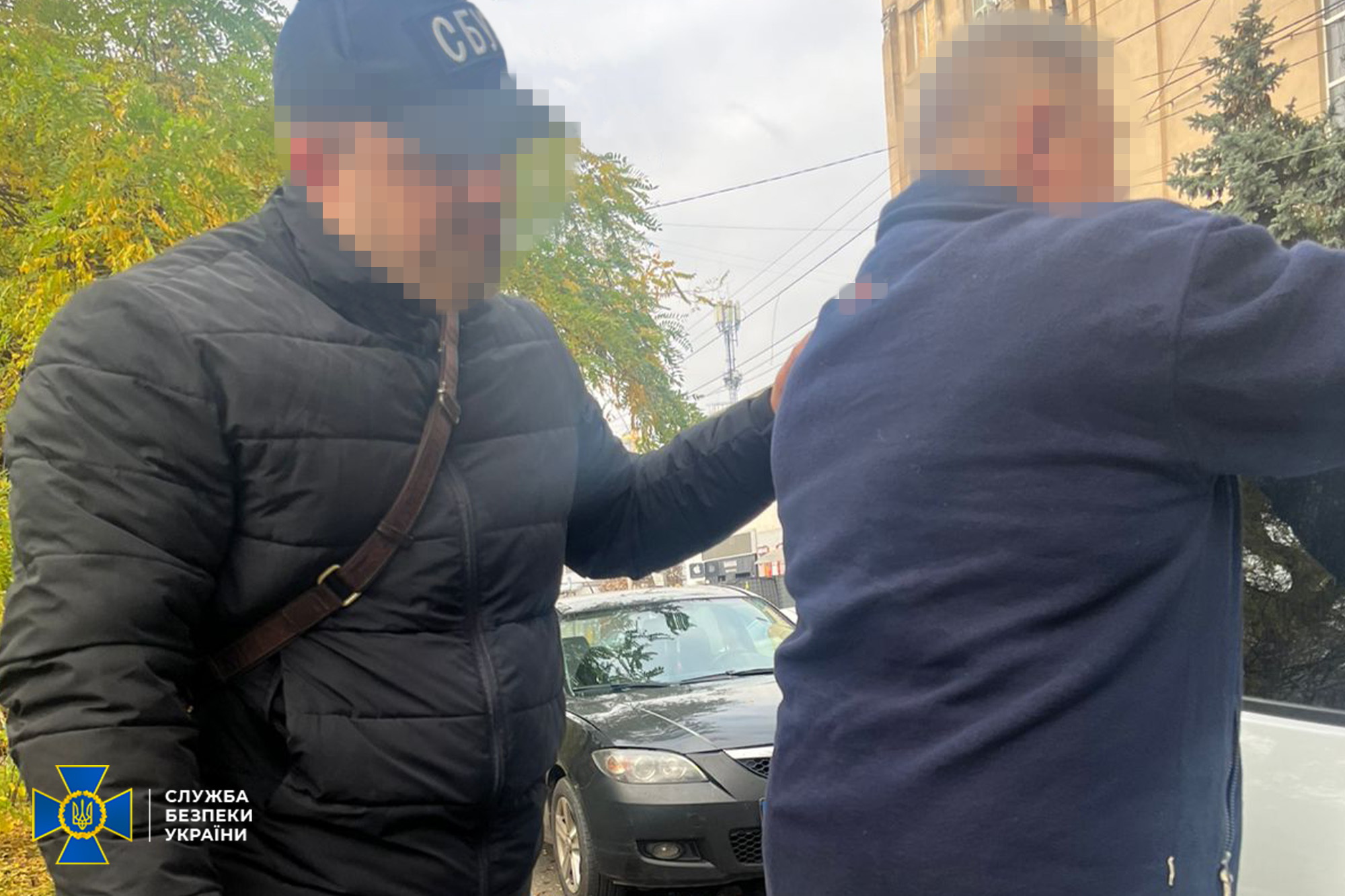 Співробітник Служби безпеки України (СБУ) у чорній куртці та кепці з написом: СБУ супроводжує чоловіка в темному одязі на вулиці. Обличчя обох чоловіків заблюрені. В нижньому лівому куті розташований логотип СБУ.