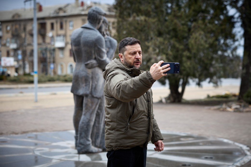 Чоловік у тактичній куртці кольору хакі стоїть на вулиці та знімає відео або робить фото на смартфон, тримаючи його перед собою. На задньому плані, у розфокусі, видно пам'ятник закоханій парі, міську забудову та дерева. Подія відбувається вдень за хмарної погоди.