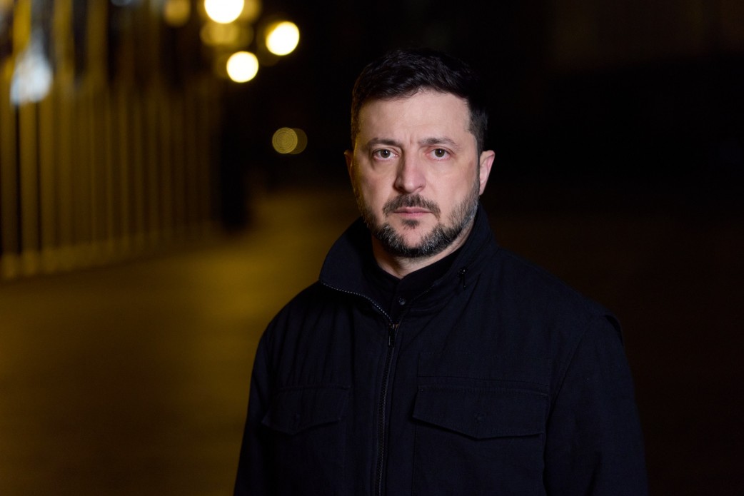 Портрет Президента України Володимира Зеленського, який стоїть на вулиці вночі, одягнений у чорну куртку, на тлі розмитих жовтих вогнів.