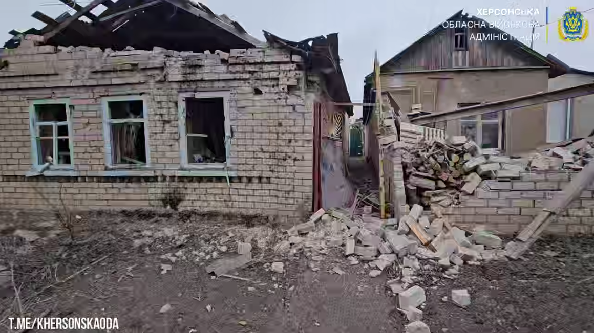 Сильно пошкоджений будинок на Херсонщині. Зруйновані цегляні стіни, розбиті вікна та відсутній дах; між двома частинами будинку лежать купи будівельного сміття. У правому верхньому куті логотип і напис: Херсонська обласна військова адміністрація.