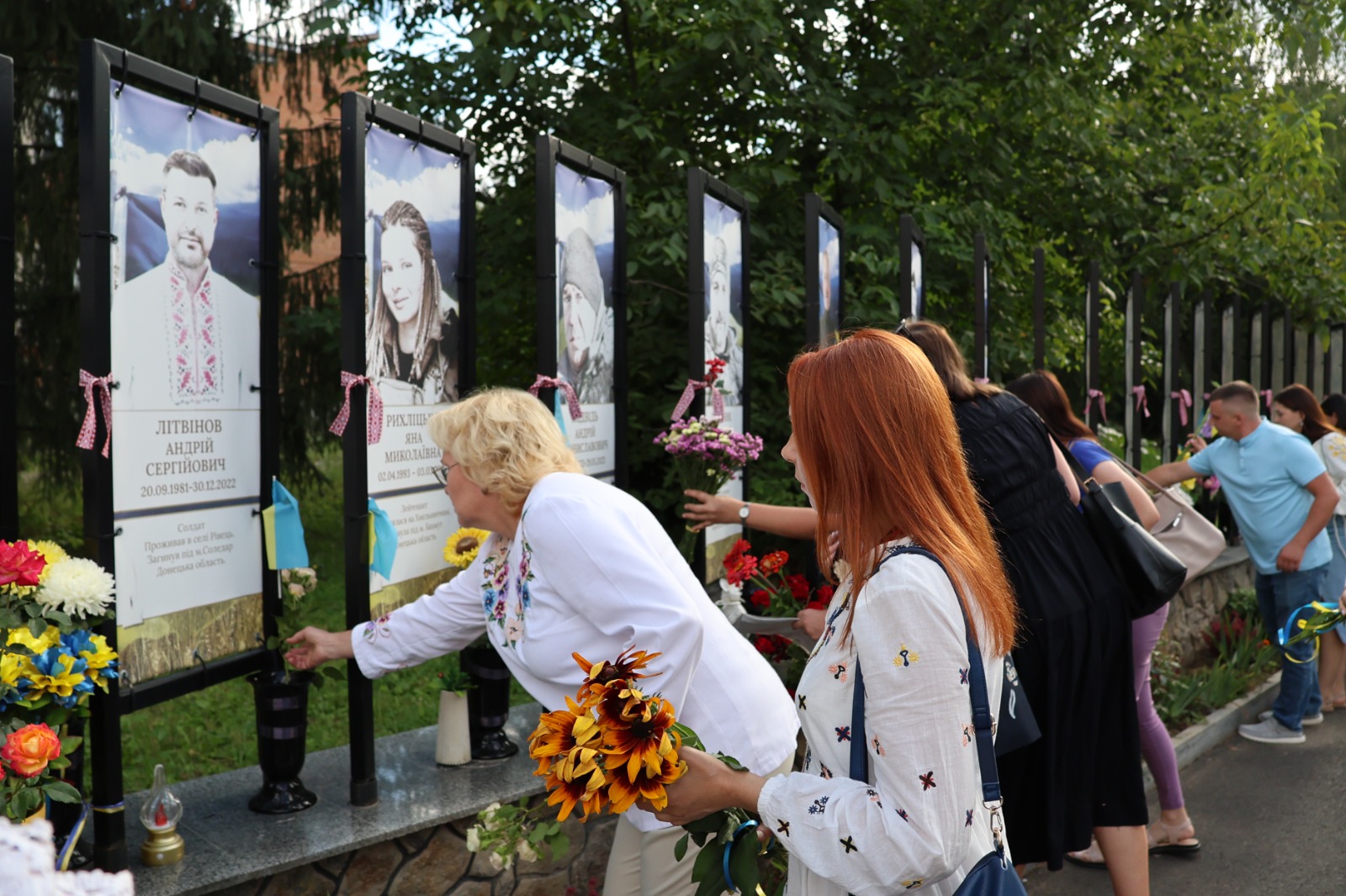 Жінки у вишиванках покладають квіти до алеї пам'яті полеглих українських воїнів. На передньому плані жінка у світлій вишитій сорочці ставить соняшники до вази біля меморіального стенда солдата Андрія Літвінова, а рудоволоса дівчина поруч тримає букет польових квітів. Вздовж алеї встановлено портрети загиблих захисників і захисниць із траурними стрічками, до яких підходять місцеві жителі.