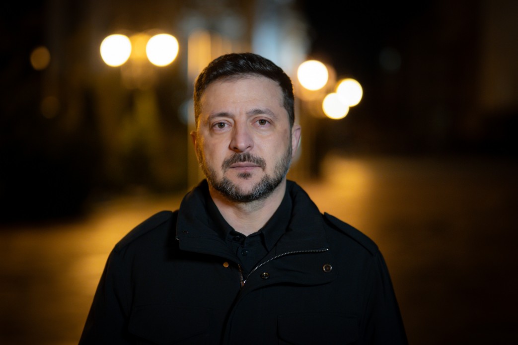 Чоловік у темному верхньому одязі на фоні нічного міста. Його погляд спрямований у камеру, а позаду видніються розмиті вогні вуличних ліхтарів.