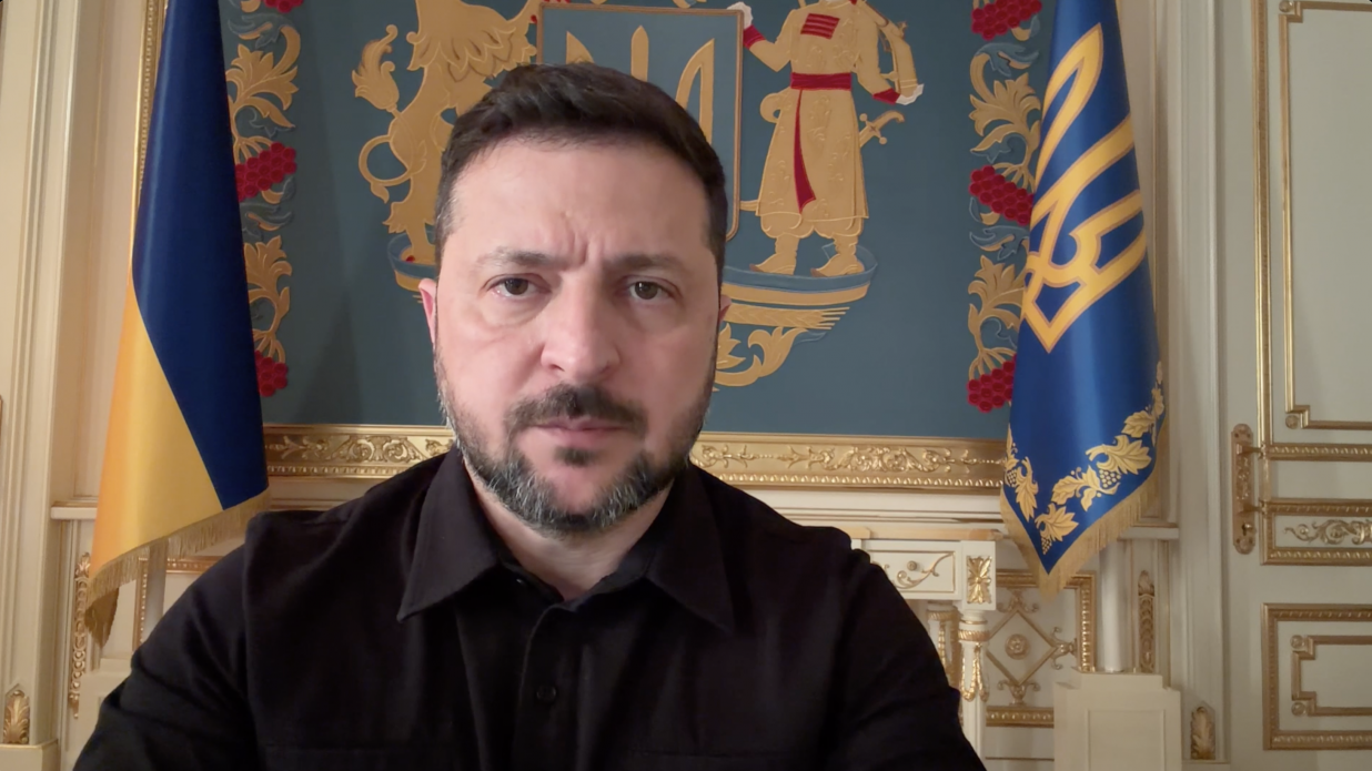 Президент України Володимир Зеленський під час офіційного звернення, зафіксований крупним планом (портрет по груди). Він одягнений у мілітарі-сорочку чорного кольору, має акуратну коротку стрижку, темну бороду та вуса. Вираз його обличчя зосереджений, серйозний та суворий; він дивиться прямо в об'єктив камери. Зйомка ведеться в офіційному інтер'єрі робочого кабінету: Ліворуч на задньому плані встановлено державний прапор України (синьо-жовте полотно із золотистою бахромою). Праворуч розташовано синій президентський штандарт із вишитим золотим тризубом по центру та рослинним золотистим орнаментом по краях. По центру безпосередньо за Президентом на стіні у золотій рамі розміщено велике художнє панно сіро-блакитного кольору із зображенням елементів Великого Державного Герба України: золотого малого герба (тризуба) на щиті, козака з мушкетом праворуч та лева ліворуч. На задньому плані також видно світлі міжкімнатні двері та стіни, оздоблені класичною золотою ліпниною і декоративними панелями.