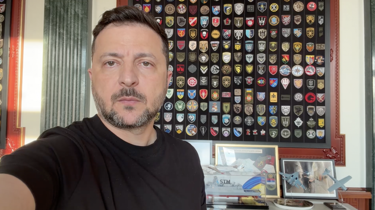 Президент України Володимир Зеленський під час офіційного відеозвернення. Він зображений великим планом на передньому плані ліворуч, одягнений у лаконічну чорну футболку. Його погляд зосереджений та спрямований прямо в камеру. На задньому плані — великий вертикальний стенд у темній дерев'яній рамі, повністю заповнений різнокольоровими шевронами та емблемами підрозділів Збройних Сил України та інших силових відомств. Під стендом на столі розташовані офіційні сувеніри та пам'ятні речі: макет військового корабля з написом «STM», невелика світлина в рамці та дві моделі літаків-винищувачів на підставках. Ліворуч видно частину зеленої інтер'єрної колони.