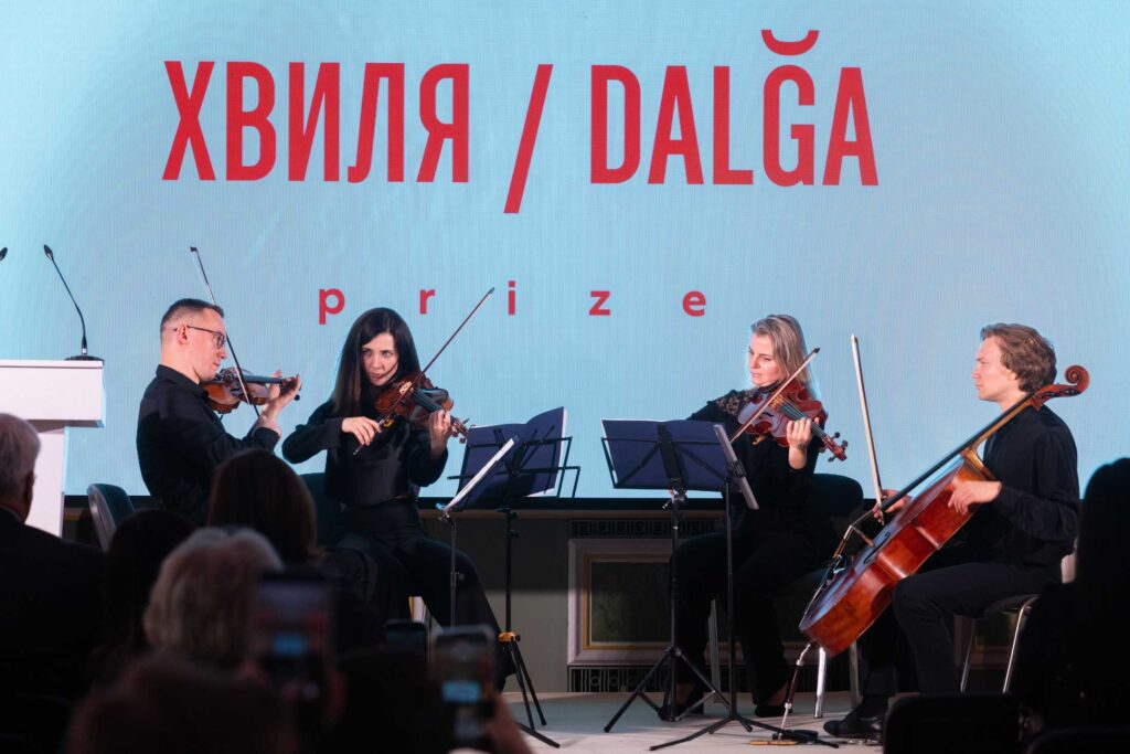 Музичний квартет у складі трьох скрипалів та віолончеліста виступає на сцені перед глядачами. На задньому плані — великий світлий екран із червоним написом «ХВИЛЯ / DALĞA prize».