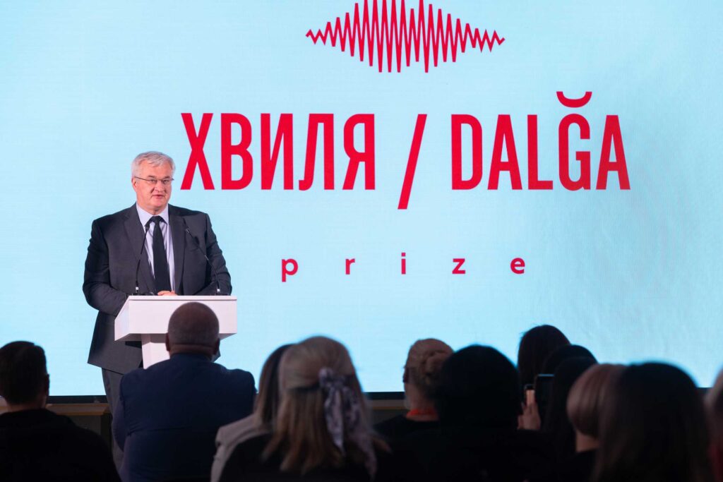 Сивочолий чоловік у класичному костюмі та окулярах виступає за білою трибуною. За його спиною — великий екран із червоним логотипом та написом «ХВИЛЯ / DALĞA prize». На передньому плані видно силуети глядачів у залі.