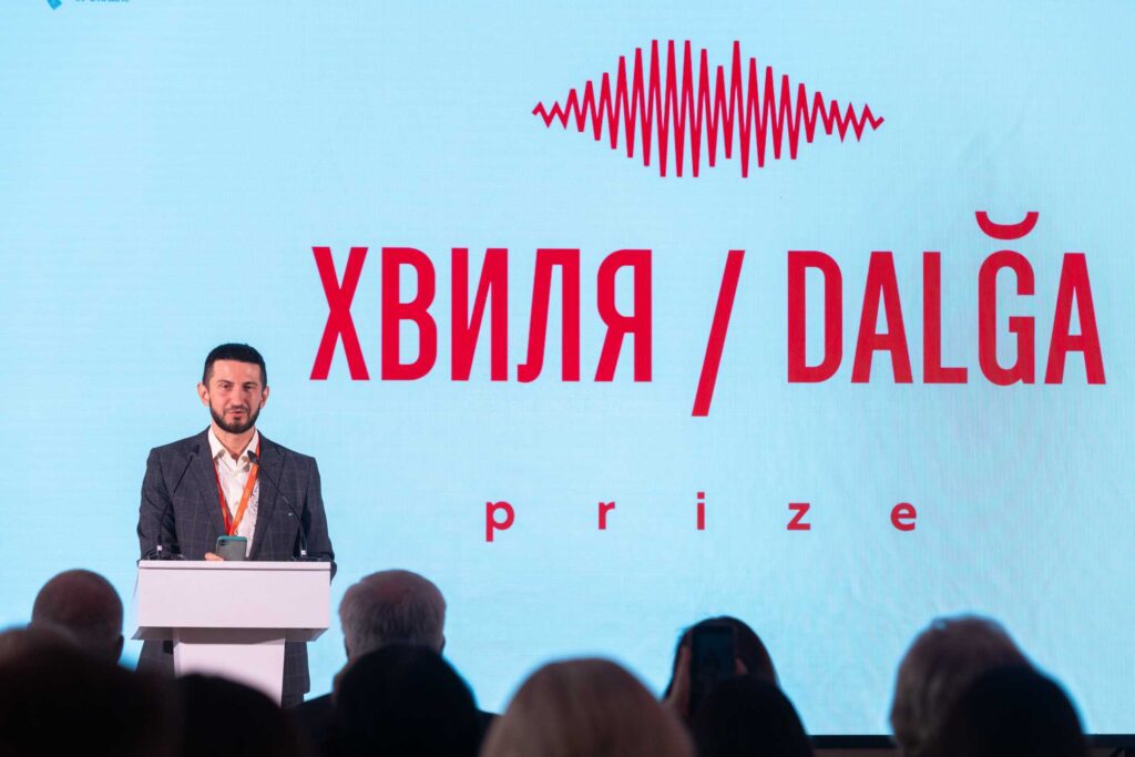 На сцені біля білої трибуни виступає чоловік із короткою темною бородою, одягнений у піджак та світлу сорочку. За його спиною — великий екран із червоним логотипом та написом «ХВИЛЯ / DALĞA prize».