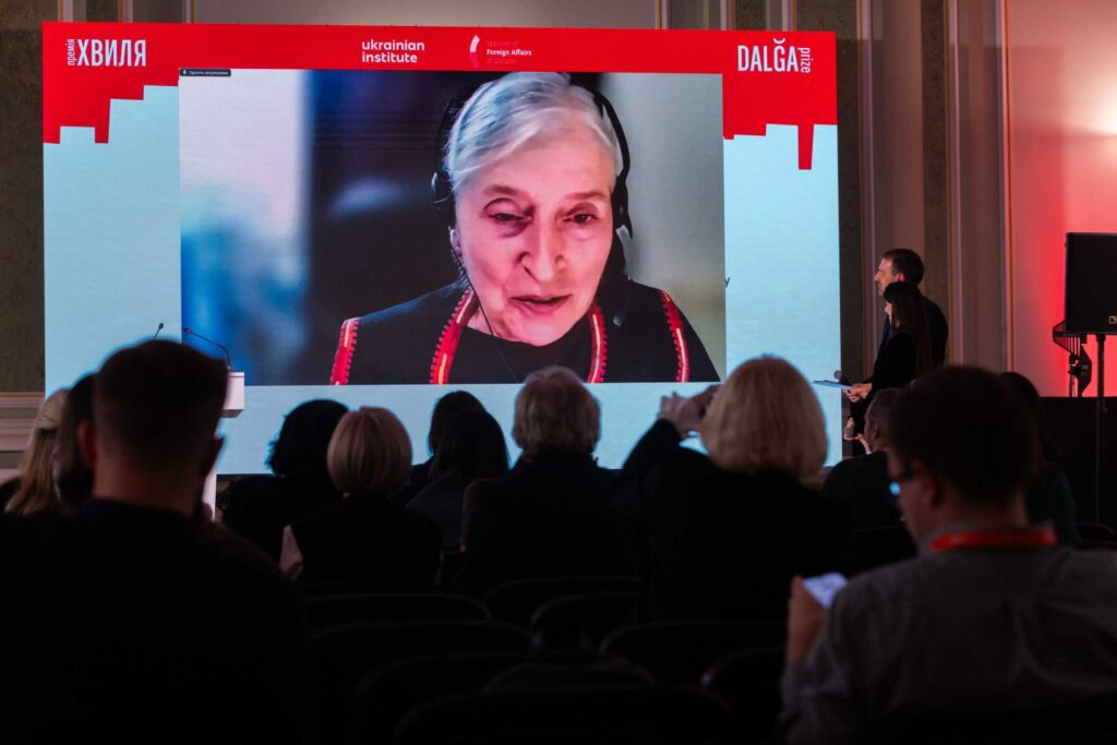 На великому екрані в залі транслюється відеозв'язок із жінкою старшого віку, яка має сиве волосся та вдягнена у навушники з гарнітурою. Глядачі в темному залі спостерігають за виступом на фоні брендованої сцени з логотипами «ХВИЛЯ» та «DALĞA prize».