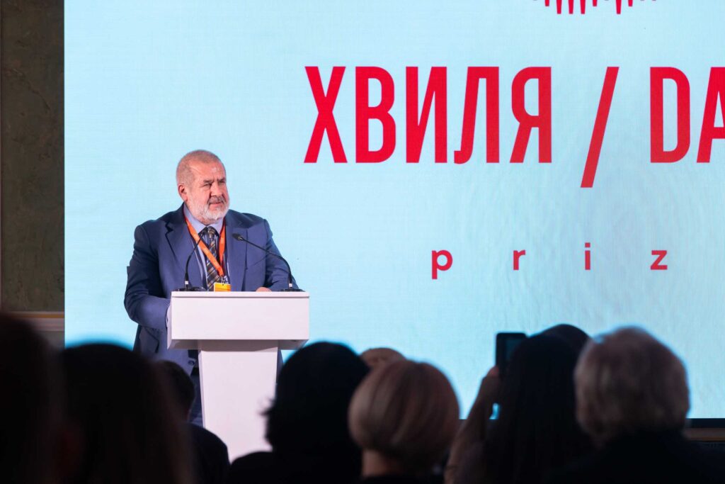 Чоловік із сивою бородою в офіційному костюмі виступає за білою трибуною з мікрофонами. За його спиною — світлий екран із великим червоним написом «ХВИЛЯ / DALĞA prize». На передньому плані видно силуети глядачів у залі.