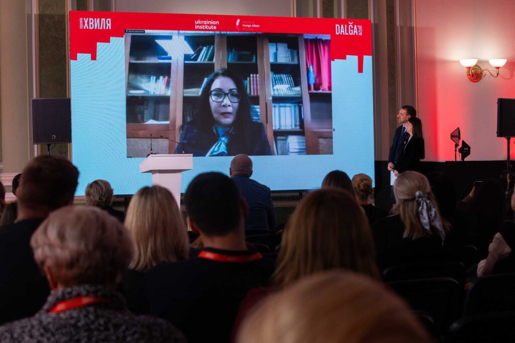 На великому екрані в залі триває онлайн-трансляція виступу жінки в окулярах на фоні книжкових полиць. Глядачі в залі спостерігають за подією, яка проходить під егідою премії «ХВИЛЯ / DALĞA prize».