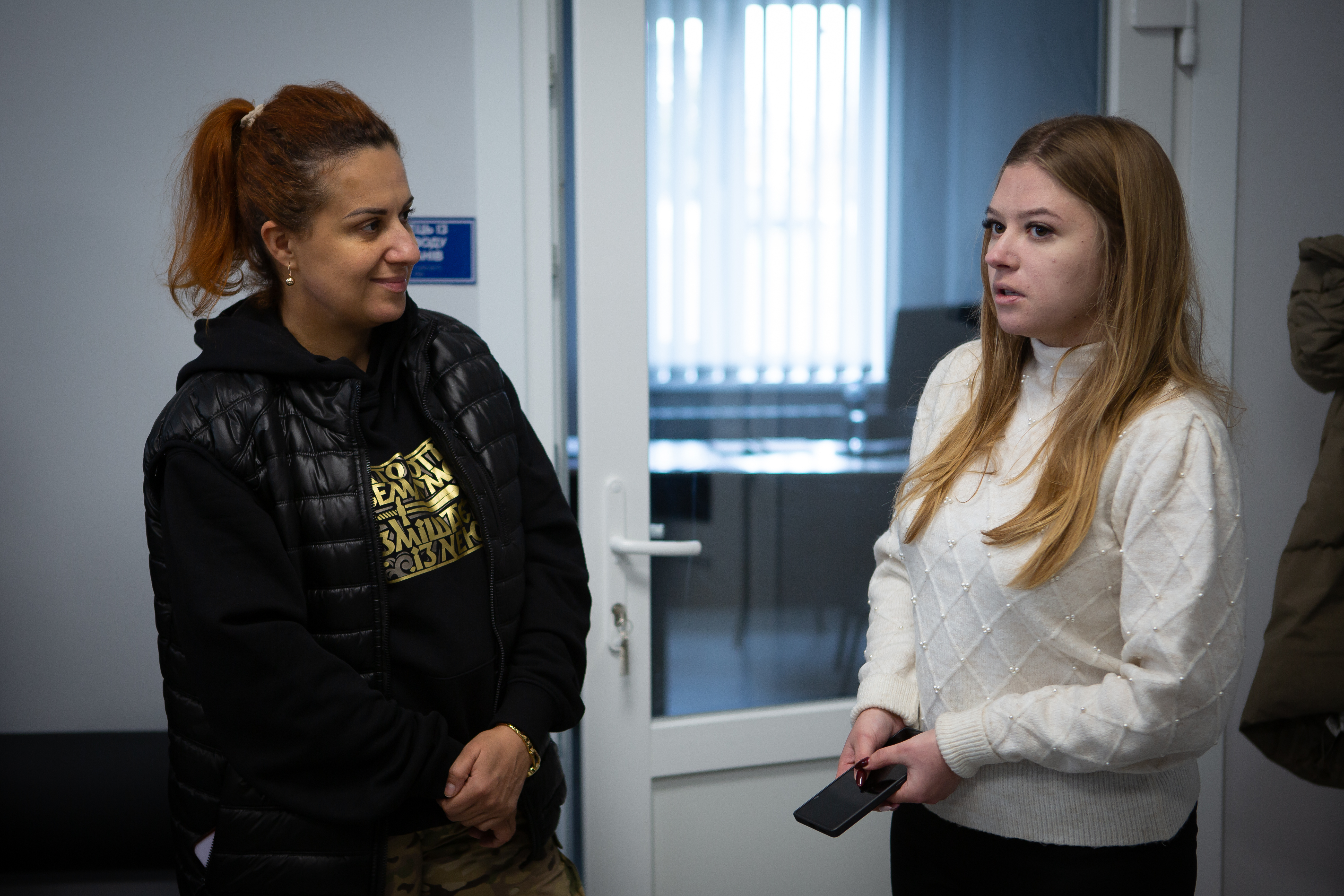 Дві жінки стоять і розмовляють у приміщенні. Жінка ліворуч, з рудим волоссям, зібраним у хвіст, одягнена у чорну стьобану жилетку та світшот, усміхається. Жінка праворуч, зі світлим довгим волоссям, одягнена у світлий в'язаний светр з візерунком, тримає в руках мобільний телефон і уважно слухає або говорить.