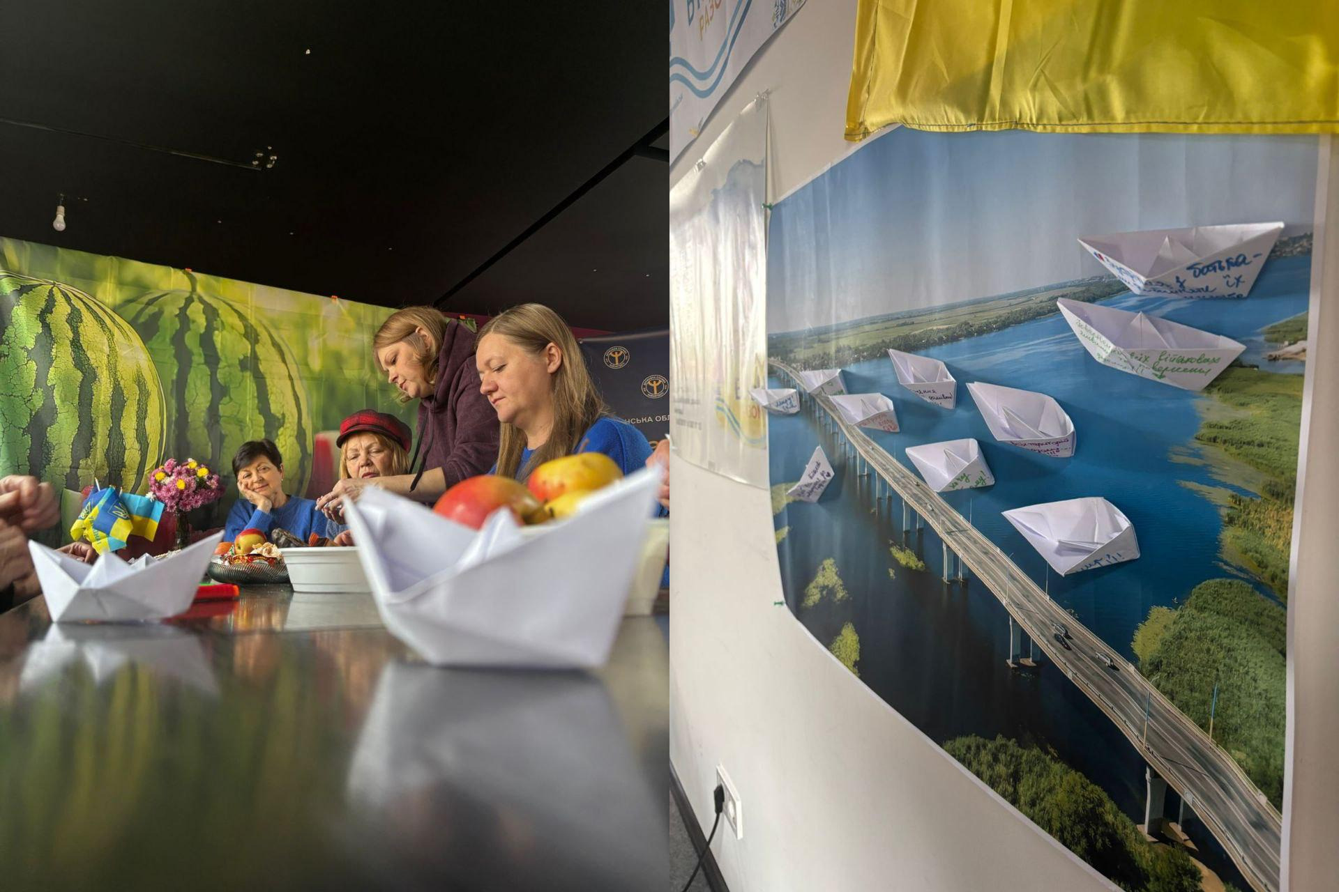 Жінки в приміщенні біля столу з фруктами та паперовими корабликами, на тлі стіни з принтом кавунів, та великий настінний плакат із зображенням мосту через річку, на який наклеєні паперові кораблики з написами.