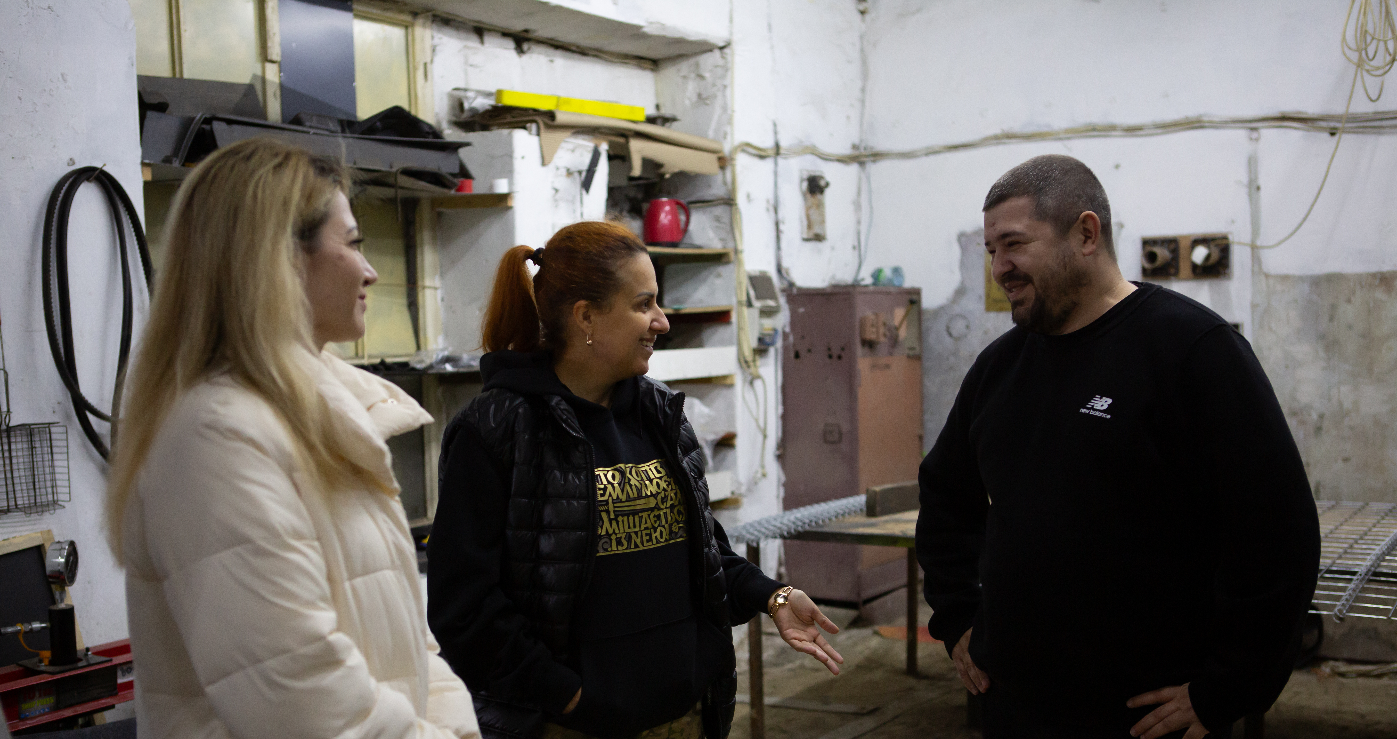 Троє людей — дві жінки та чоловік — спілкуються у, ймовірно, майстерні або виробничому приміщенні. Жінка ліворуч одягнена у світлу куртку, жінка в центрі — у чорну жилетку та світшот, а чоловік праворуч — у чорний светр. Вони посміхаються та жестикулюють. На задньому плані видно полиці, обладнання та необроблені білі стіни.