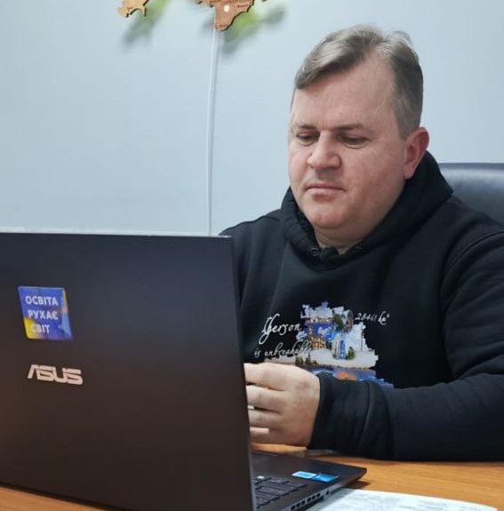 Чоловік у чорному худі із патріотичним принтом сидить за столом перед відкритим ноутбуком. На кришці ноутбука наклеєно стікер із написом «Освіта рухає світ», а на стіні позаду видніється частина дерев'яної мапи України.