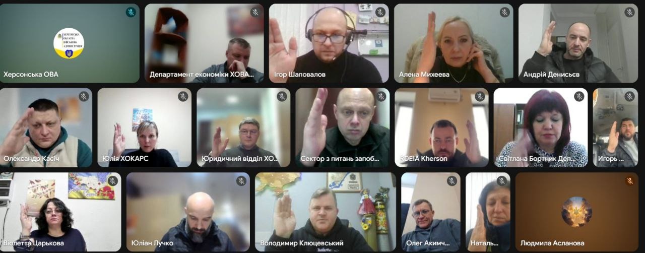 Скриншот онлайн-наради у форматі відеоконференції з багатьма учасниками, які одночасно тримають піднятою одну руку. У кутку екрана розміщено логотип Херсонської ОВА.