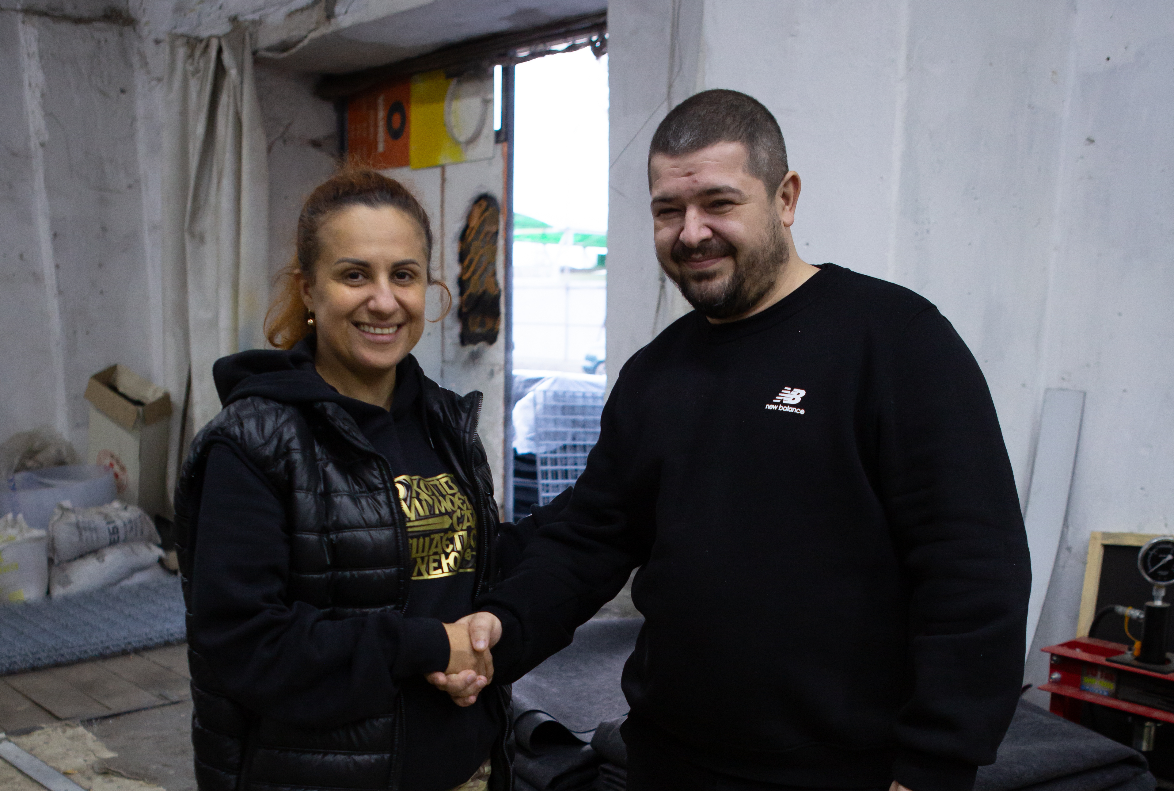 Жінка та чоловік потискають один одному руки, стоячи у приміщенні, схожому на майстерню або склад. Обидва посміхаються. Жінка одягнена у чорну стьобану жилетку та світшот, чоловік — у чорний светр. На задньому плані видно відкритий дверний проріз, через який проникає денне світло, та мішки і предмети, що стоять усередині.