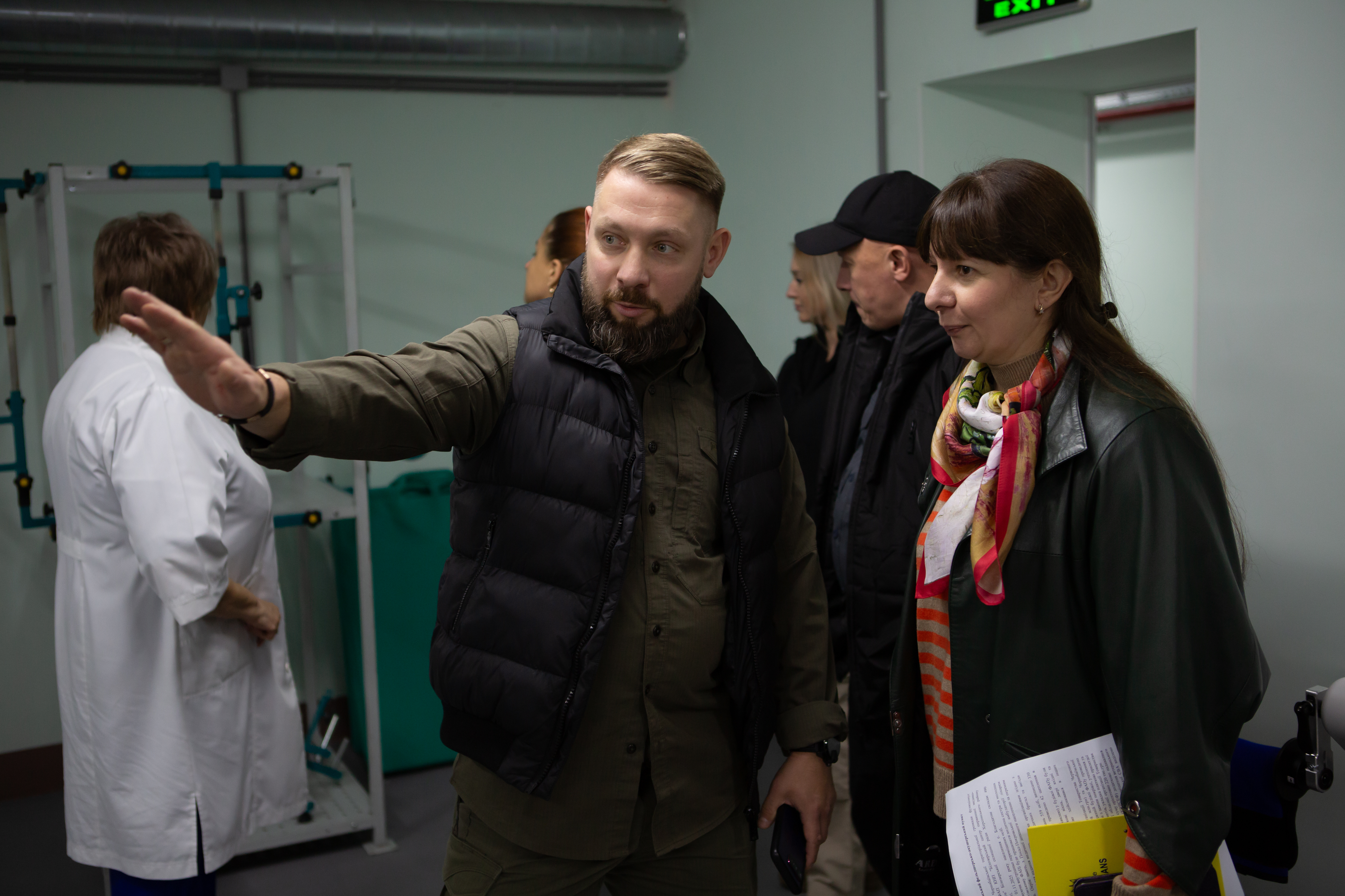 Чоловік у стьобаному чорному жилеті та сорочці кольору хакі жестом руки щось пояснює жінці, яка стоїть праворуч від нього і тримає документи. На ній шкіряна куртка та яскравий шарф. На задньому плані видно ще кілька людей, а також медичне або реабілітаційне обладнання (скляна конструкція та жінка в білому халаті), що свідчить про перебування у медичному закладі.