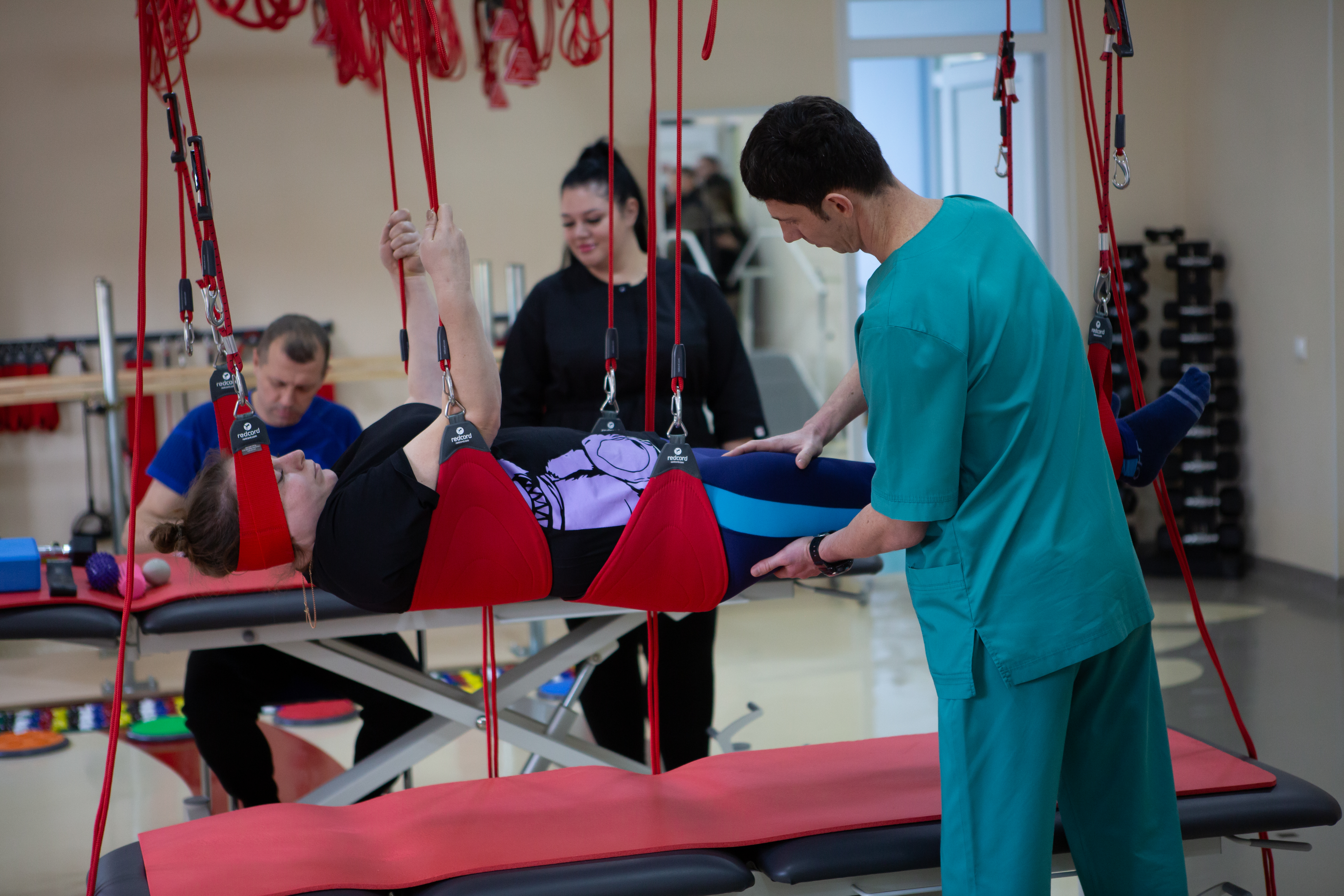 Сцена фізичної реабілітації. Жінка лежить на спині на терапевтичному столі, її тіло частково підтримується червоними стропами під час кінезіотерапії. Фізіотерапевт у бірюзовому медичному костюмі стоїть праворуч і контролює її ногу. На задньому плані видно ще одного пацієнта, що сидить за столом, та інструктора, які спостерігають за процесом.