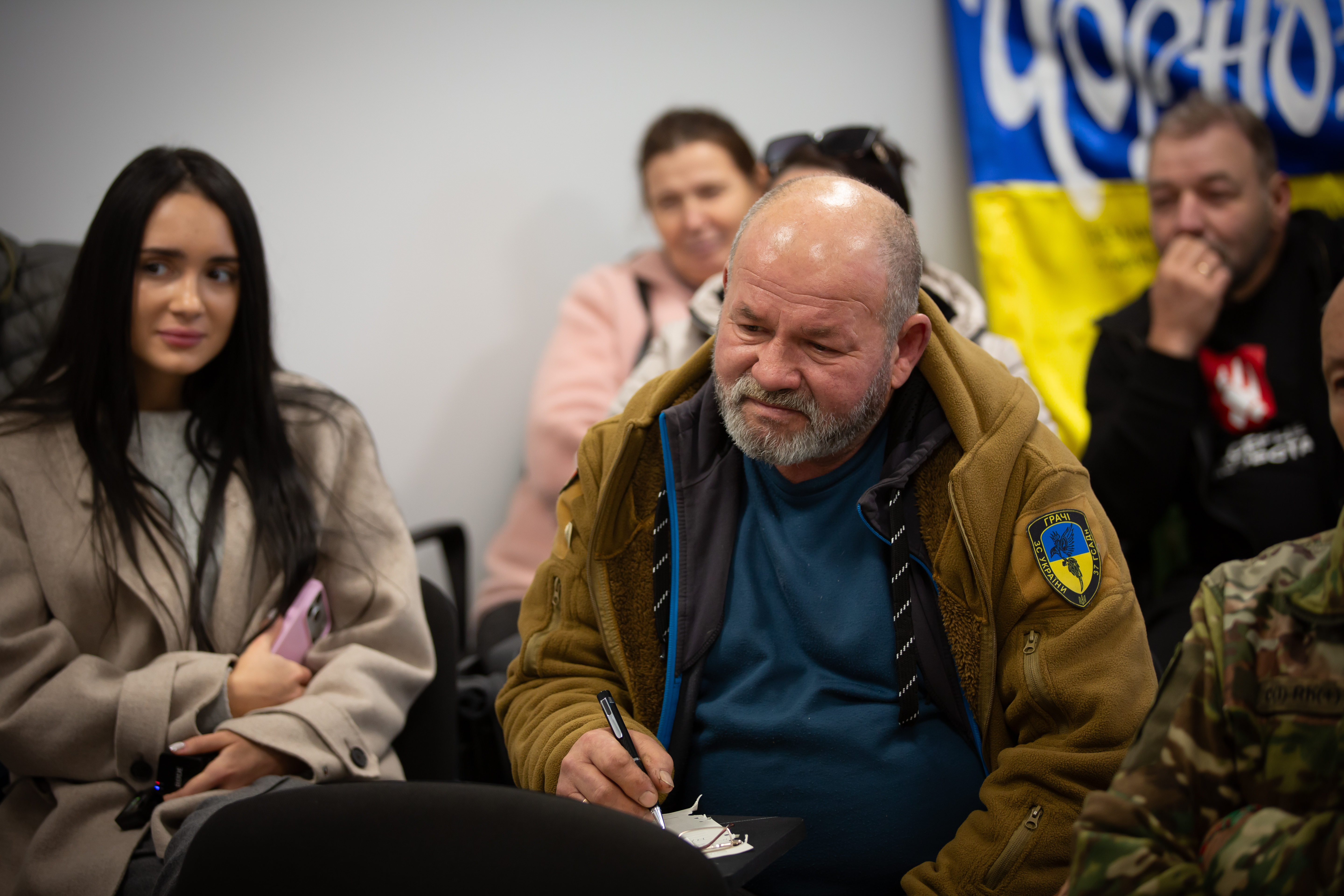 Чоловік середнього віку з бородою та лисиною, одягнений у теплу світло-коричневу куртку із шевроном, сидить і щось записує ручкою на невеликому блокноті. Ліворуч від нього сидить молода жінка з довгим темним волоссям у світлому пальті. На задньому плані видно інших учасників зустрічі, зокрема, прапор України та синій прапор із написом.