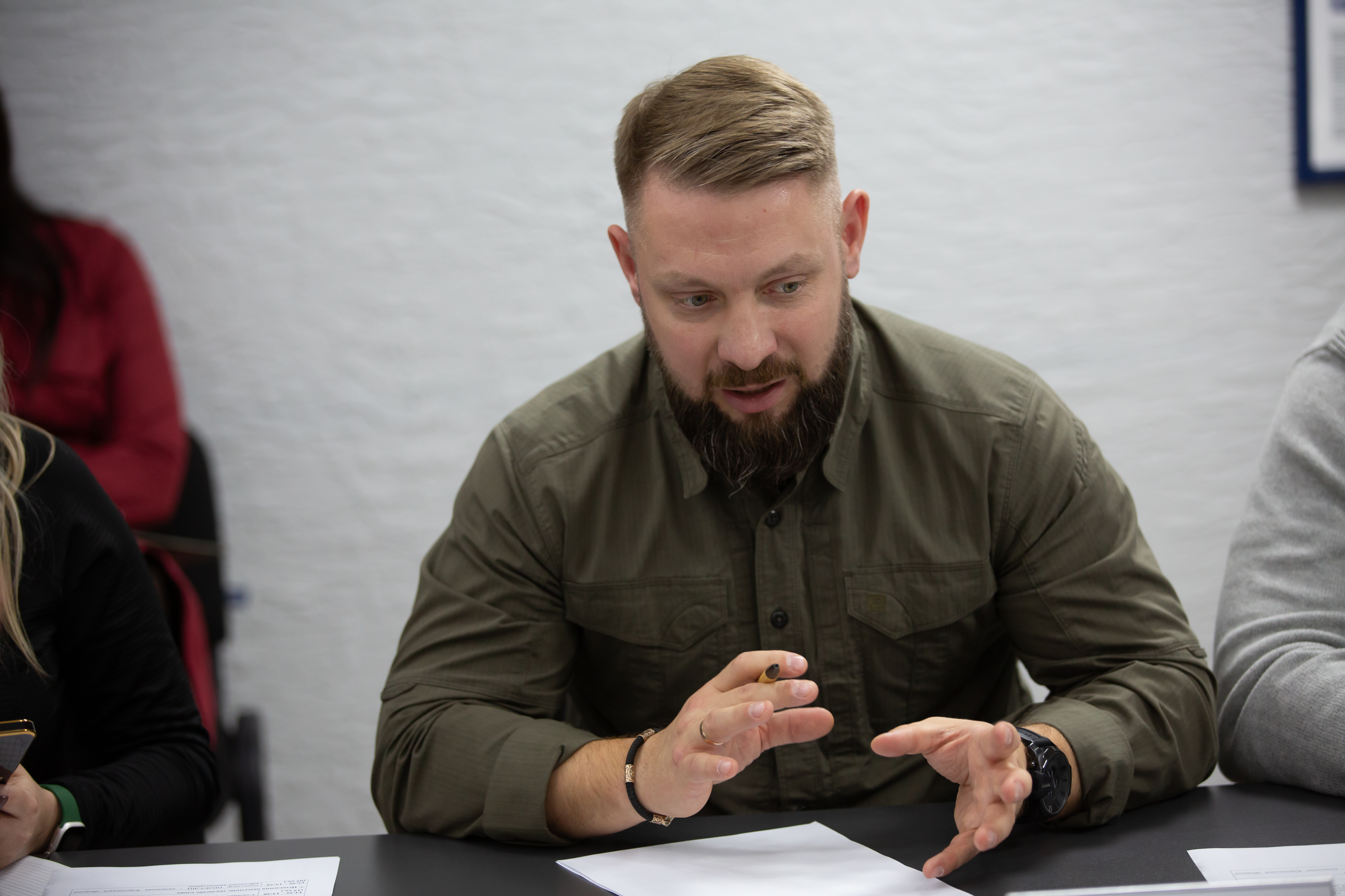 Бородатий чоловік середнього віку з коротко стриженим волоссям сидить за столом під час наради. Він одягнений у сорочку кольору хакі, активно жестикулює руками над паперами, тримаючи в правій руці ручку. Навколо нього частково видно інших учасників зустрічі.