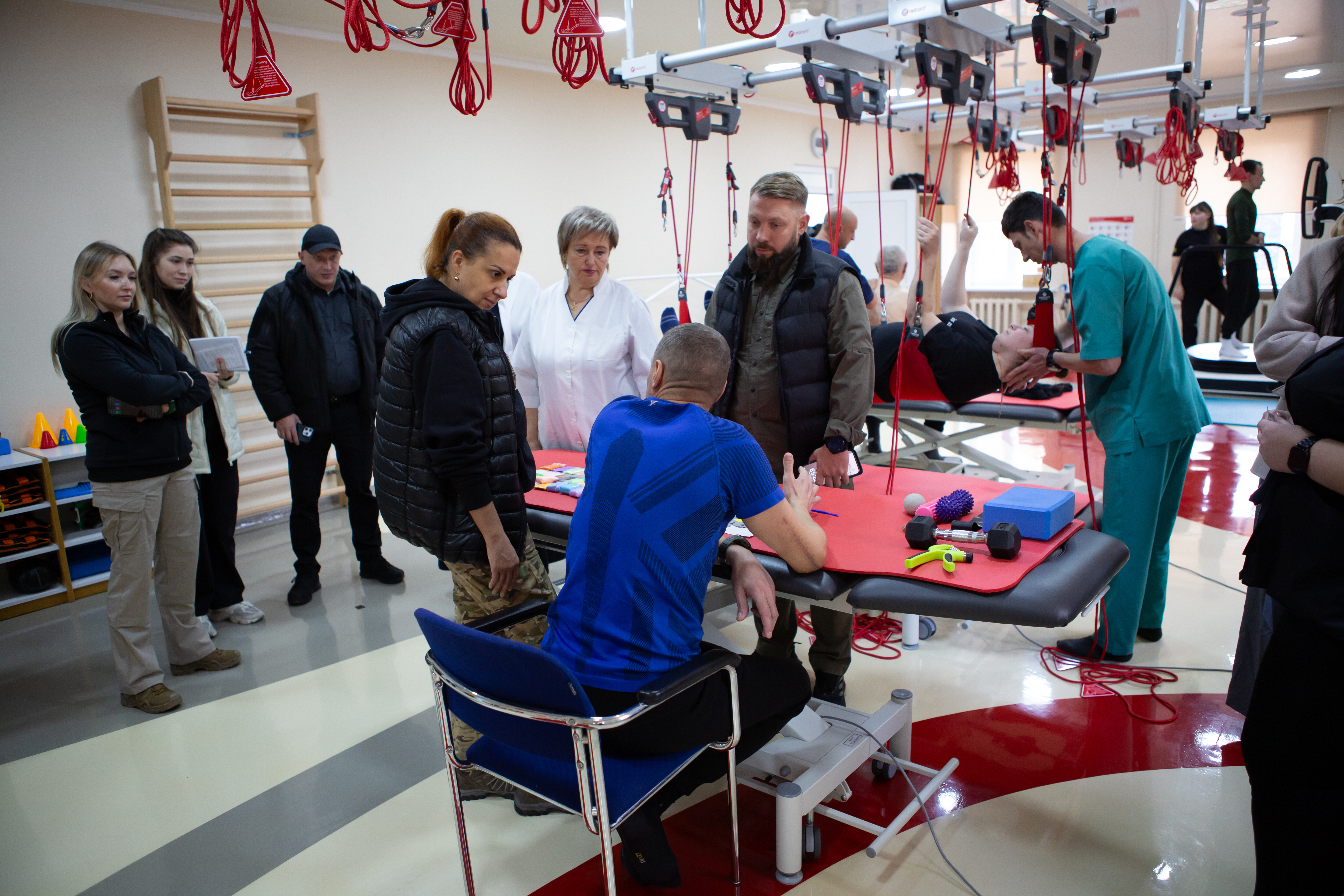 Група людей, включаючи офіційних осіб та медичний персонал, відвідує реабілітаційний центр. У центрі уваги — пацієнт у синій футболці, який сидить перед столом, спілкуючись із жінкою у чорній жилетці та чоловіком у стьобаному жилеті. На задньому плані інший пацієнт проходить реабілітацію на спеціальному обладнанні з червоними стропами під наглядом фізіотерапевта.