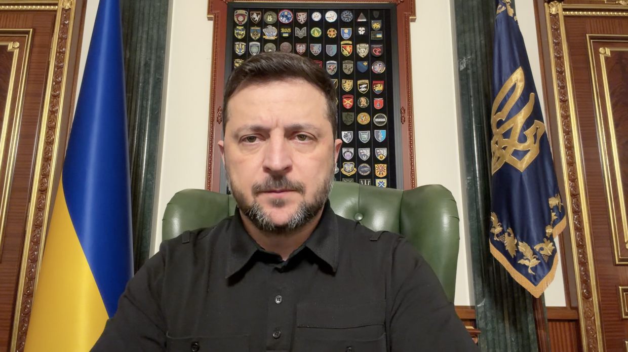 Чоловік у чорній сорочці сидить у кабінеті на фоні державного прапора України та прапора з гербом. Позаду нього на стіні розміщена дошка з численними військовими шевронами.