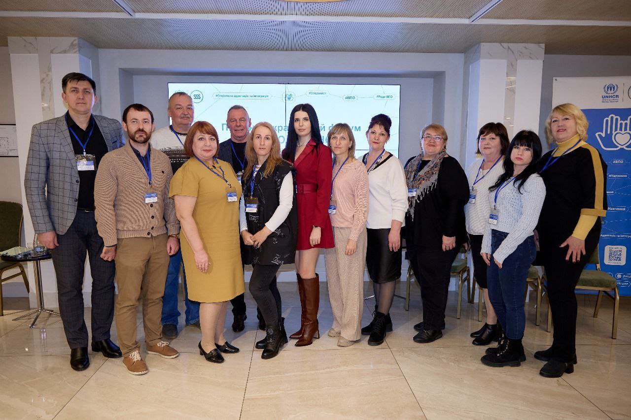 Групове фото, зроблене в приміщенні, на якому зображено дванадцять осіб (чоловіків і жінок різного віку) у повсякденному та діловому одязі, що стоять у ряд, учасники форуму. На задньому плані видно банер з логотипами та написом: UNHCR (УВКБ ООН).