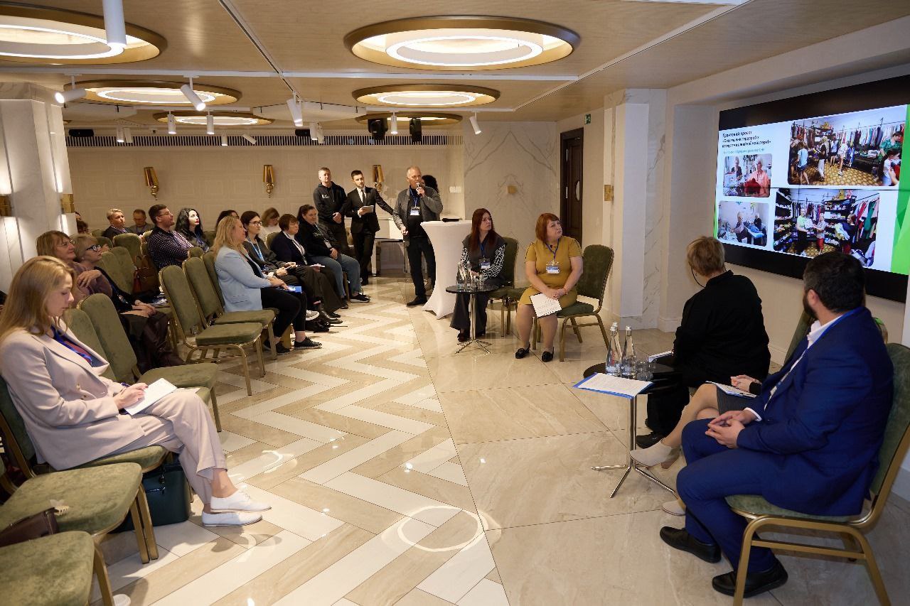 Вид на конференц-зал під час форуму, де близько 20-30 учасників сидять на зелених стільцях, слухаючи доповідача, що стоїть біля екрана. На великому екрані праворуч відображаються фотографії одягу та інтер'єру.