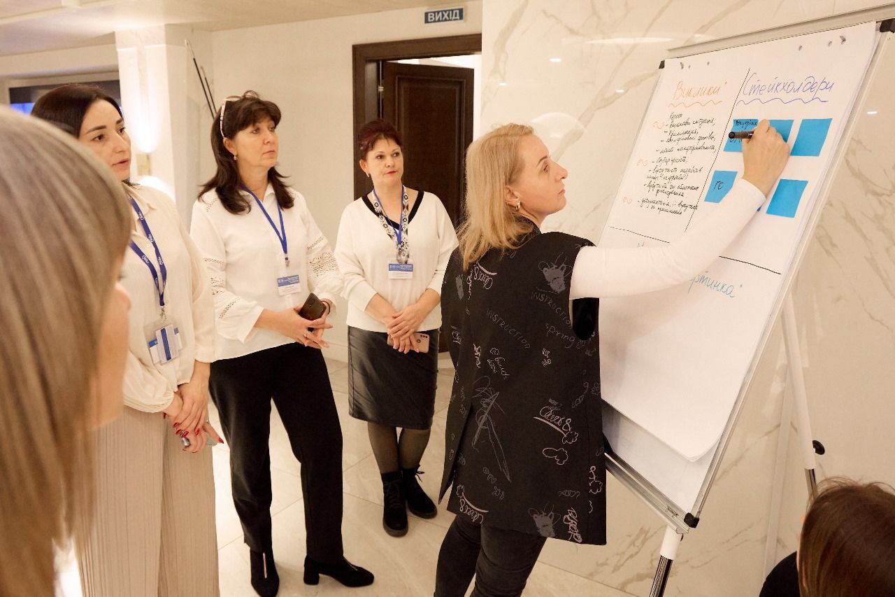 Чотири жінки на форумі. Одна жінка, одягнена у чорно-білий жилет, пише маркером на фліпчарті, а три інші жінки уважно спостерігають. На фліпчарті видно українські написи: Виклики та Стейкхолдери.