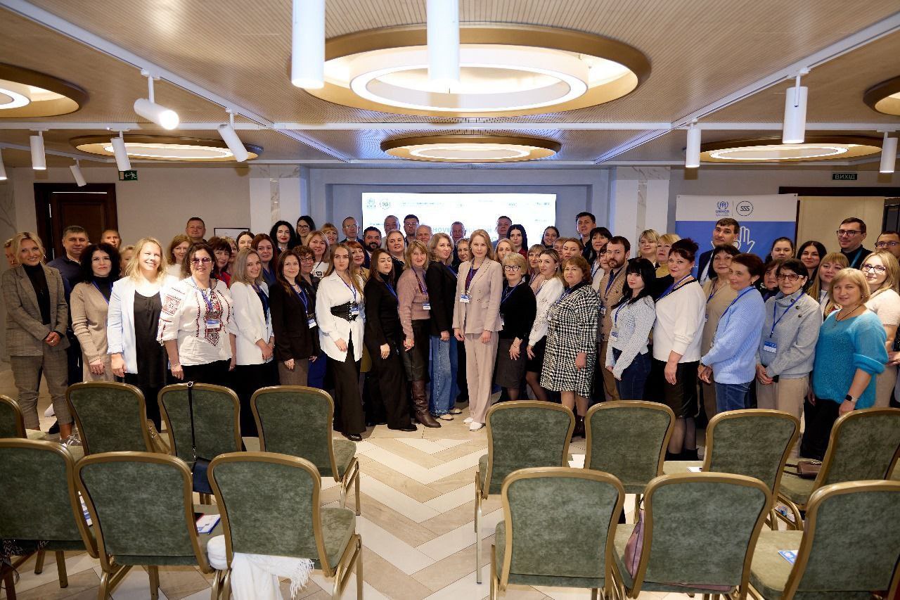 Велике групове фото, зроблене у конференц-залі з рядами порожніх зелених стільців на передньому плані. Велика група учасників (близько 50-60 осіб) стоїть ззаду, посміхаючись. На задньому плані над головами видно екран з логотипами: та таблички ВИХІД.