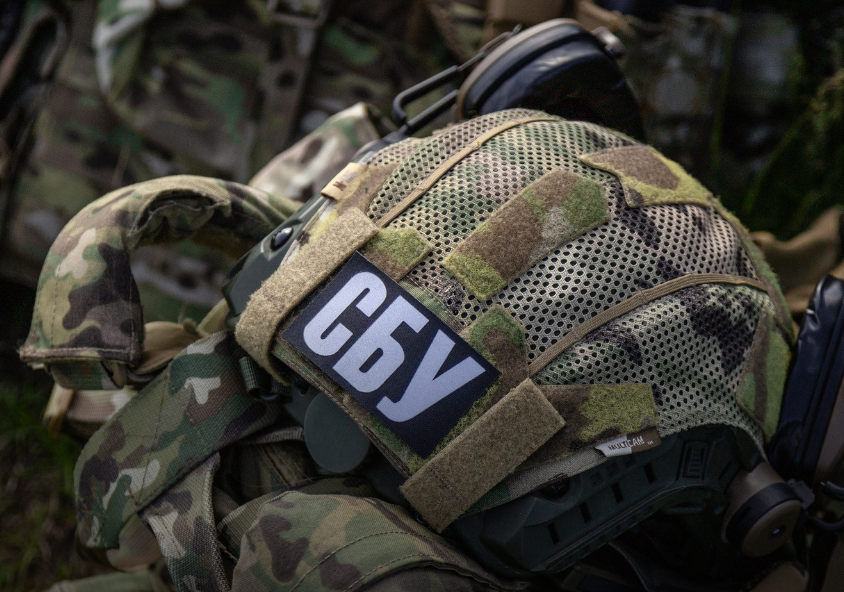 Військовий шолом у камуфляжному чохлі з нашивкою «СБУ» лежить поверх іншого тактичного спорядження. До шолома прикріплені активні навушники.