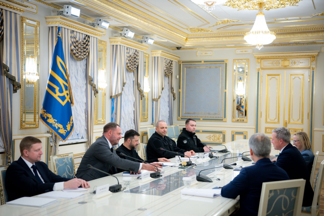 Зустріч у розкішному залі, де Президент України Володимир Зеленський та його команда сидять за довгим столом навпроти іноземної делегації. На столі лежать документи, а позаду — прапор із Тризубом.