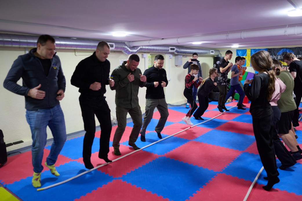Група дорослих чоловіків (включно з Президентом Зеленським) та підлітків бере участь у спортивній активності, перестрибуючи через натягнуту мотузку. Вони знаходяться у спортивному залі з килимком із синіх та червоних квадратів.