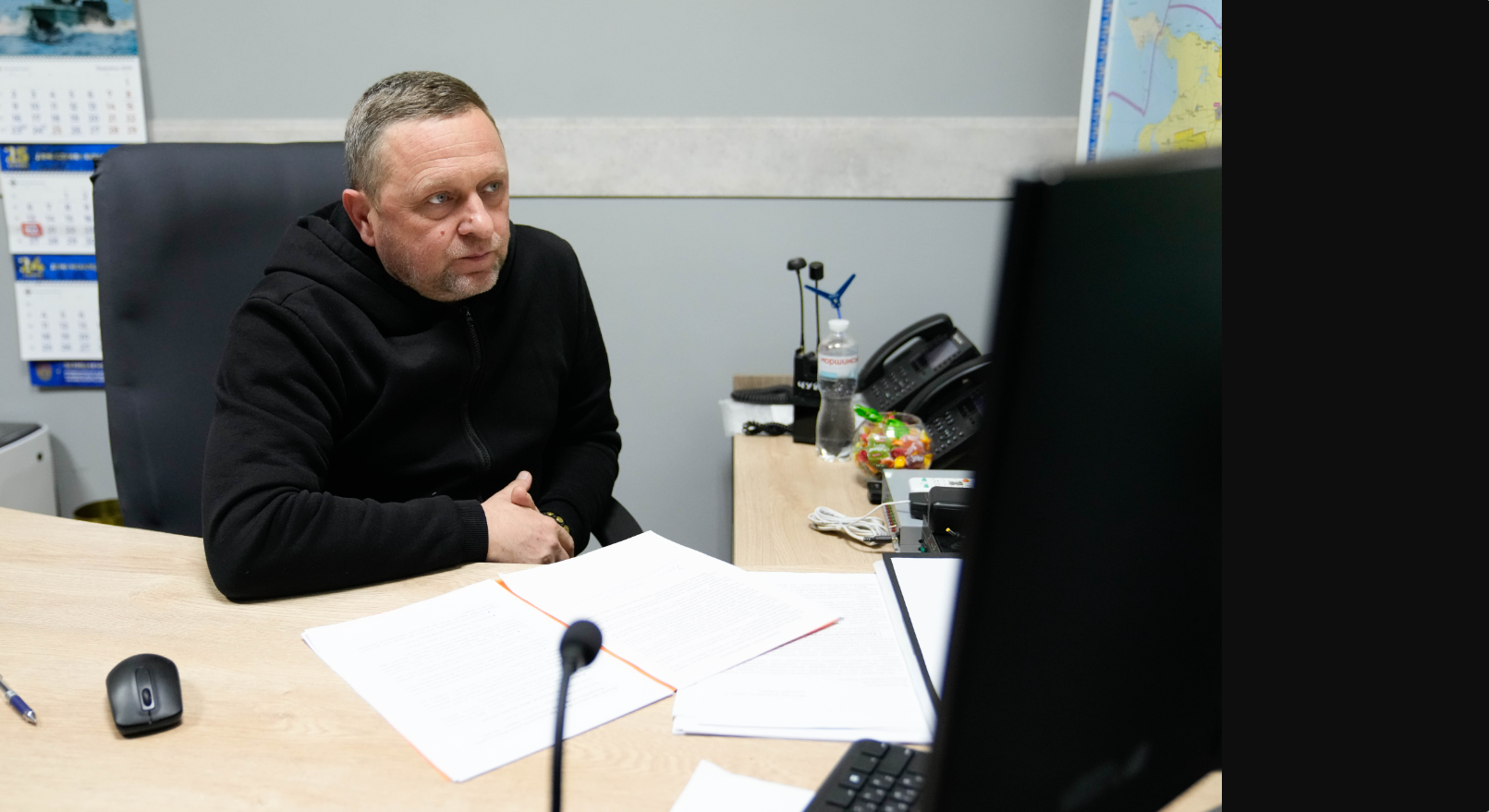 Чоловік у чорному худі сидить за робочим столом перед монітором комп'ютера. На столі лежать відкриті документи, стоїть мікрофон та офісне приладдя. На стіні позаду видно частину календаря та карти.