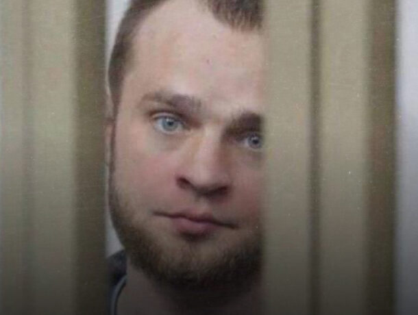 Крупний план обличчя світлошкірого чоловіка з блакитними очима та бородою, знятий через вертикальні щілини, схожі на ґрати або дерев'яні планки, які приховують частину обличчя.