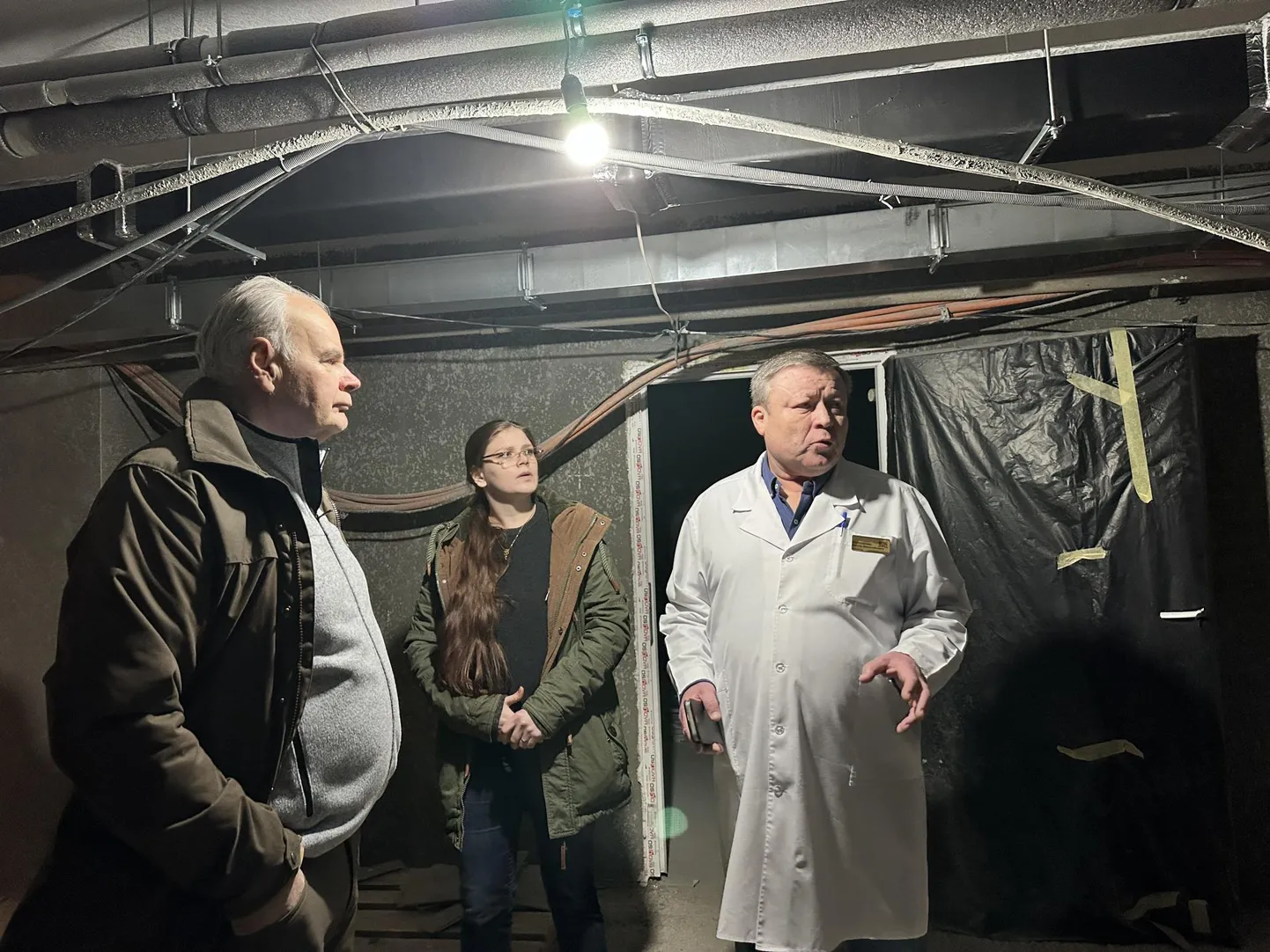 Чоловік у медичному халаті спілкується з двома людьми у підвальному приміщенні з відкритими комунікаціями на стелі.