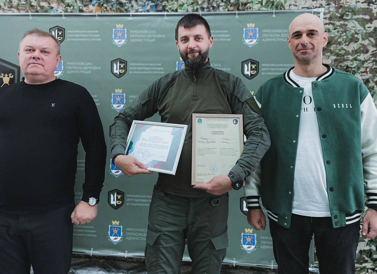 Троє чоловіків стоять разом: чоловік у військовому одязі з бородою в центрі тримає дві рамки з сертифікатами чи нагородами, його оточують двоє чоловіків у цивільному одязі.