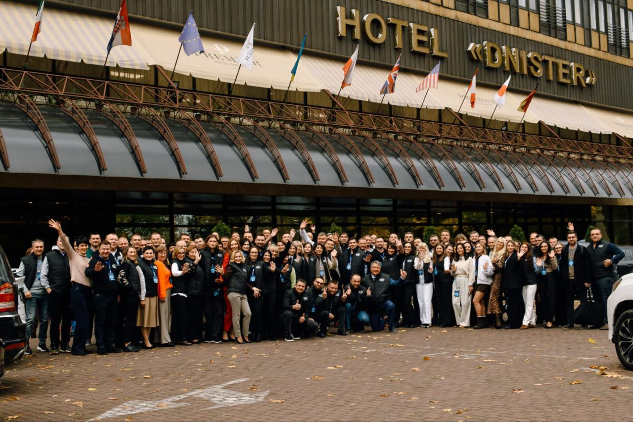 Велика група людей у переважно чорному та діловому одязі, багато хто з яких усміхається та махає руками, стоїть на тротуарі для спільного фото на тлі входу в готель Дністер (HOTEL DNISTER). Над входом майорять прапори різних країн та міжнародних організацій.