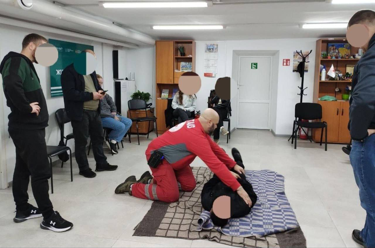 Інструктор у червоно-сірій формі (Перша Допомога) стоїть на колінах і перевертає особу або манекен, що лежить на ковдрі на підлозі, демонструючи техніку переведення у стабільне бокове положення під час курсу першої допомоги. Кілька учасників спостерігають за демонстрацією, стоячи або сидячи навколо.