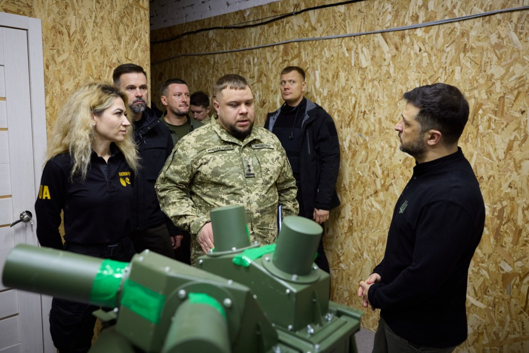  Президент України Володимир Зеленський (в чорній кофті) спілкується з групою військових та цивільних осіб у приміщенні, обшитому OSB-плитами. На передньому плані видно частину військового обладнання зеленого кольору. Чоловік у центрі, одягнений у піксельну військову форму, розповідає про обладнання.