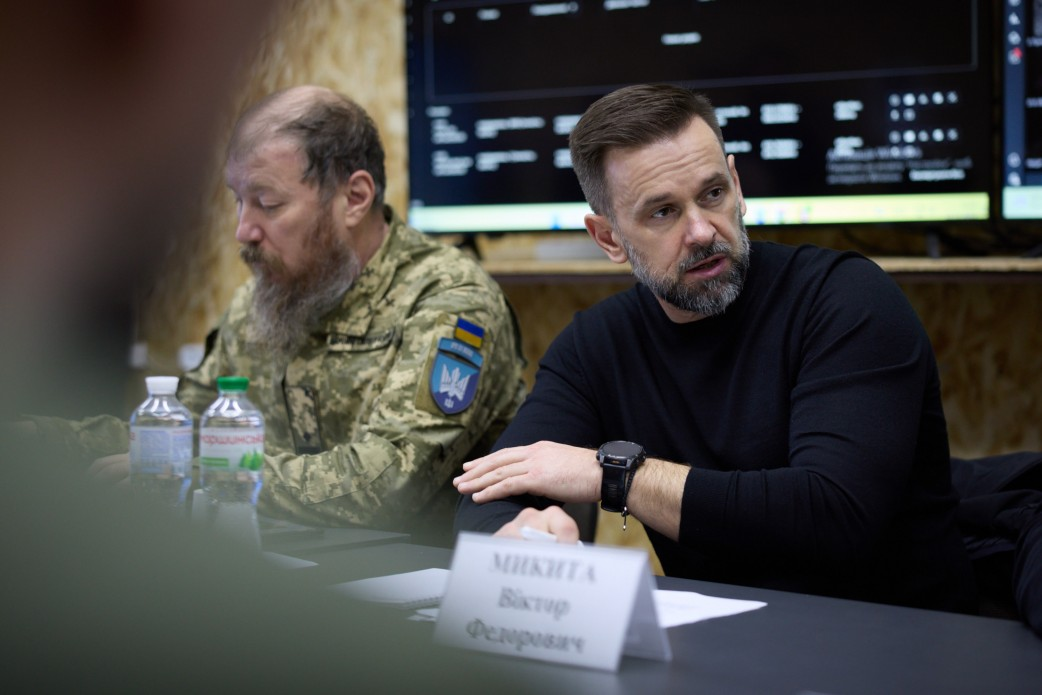 Крупний план двох чоловіків за столом під час наради. На передньому плані — чоловік у чорному светрі, який жестикулює. Поруч сидить чоловік у військовій формі з довгою бородою. На столі видно пляшки з водою, а на задньому плані — екран.