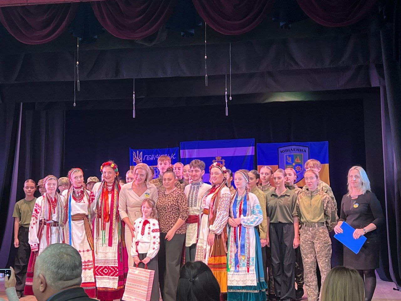 Група молодих людей та дорослих стоять на сцені. Частина молоді одягнена у традиційний український одяг (вишиванки та вінки), а частина — у військову або камуфляжну форму. В центрі стоять декілька дорослих жінок. На задньому плані видно банери в синьо-жовтих кольорах із написами, один з яких містить слово ГЕНІЧЕСЬК, а інший — ЮВІЛЕЙНА.