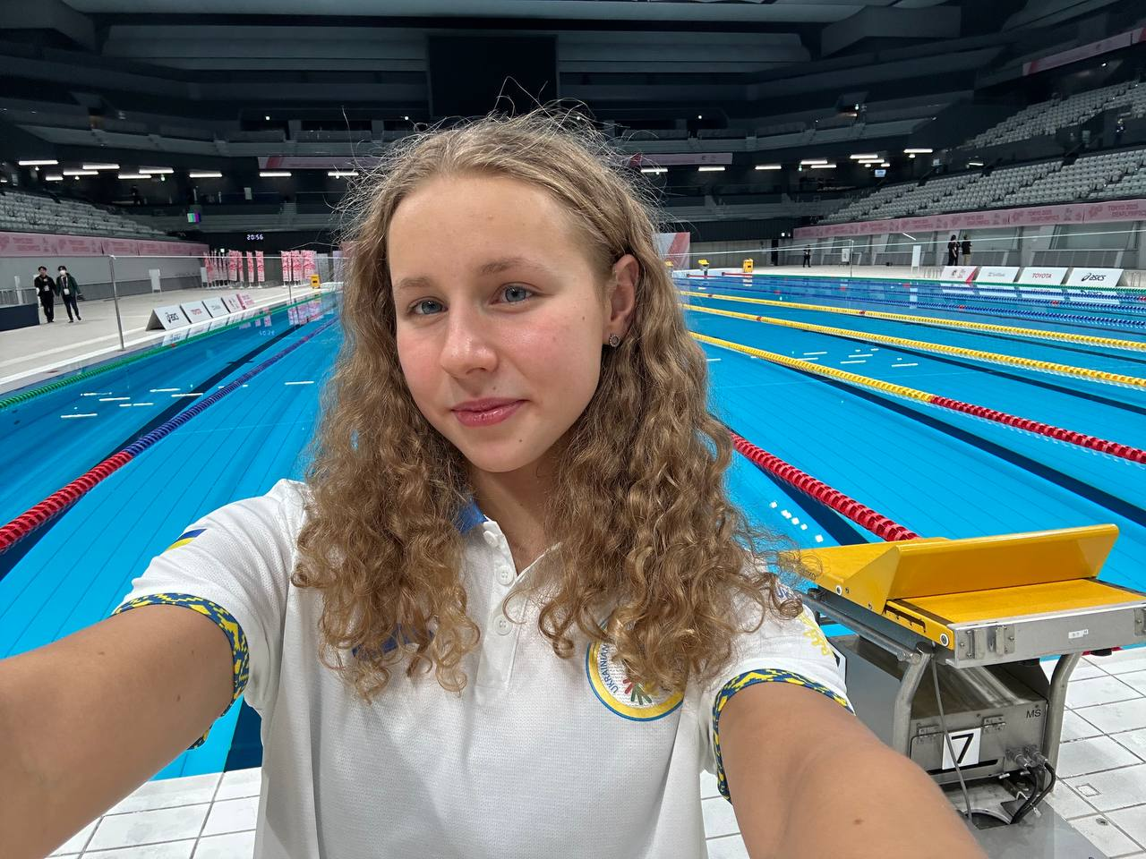 Селфі молодої спортсменки з кучерявим волоссям у білій формі з українською символікою, зроблене на тлі олімпійського басейну з доріжками та трибунами на задньому плані.