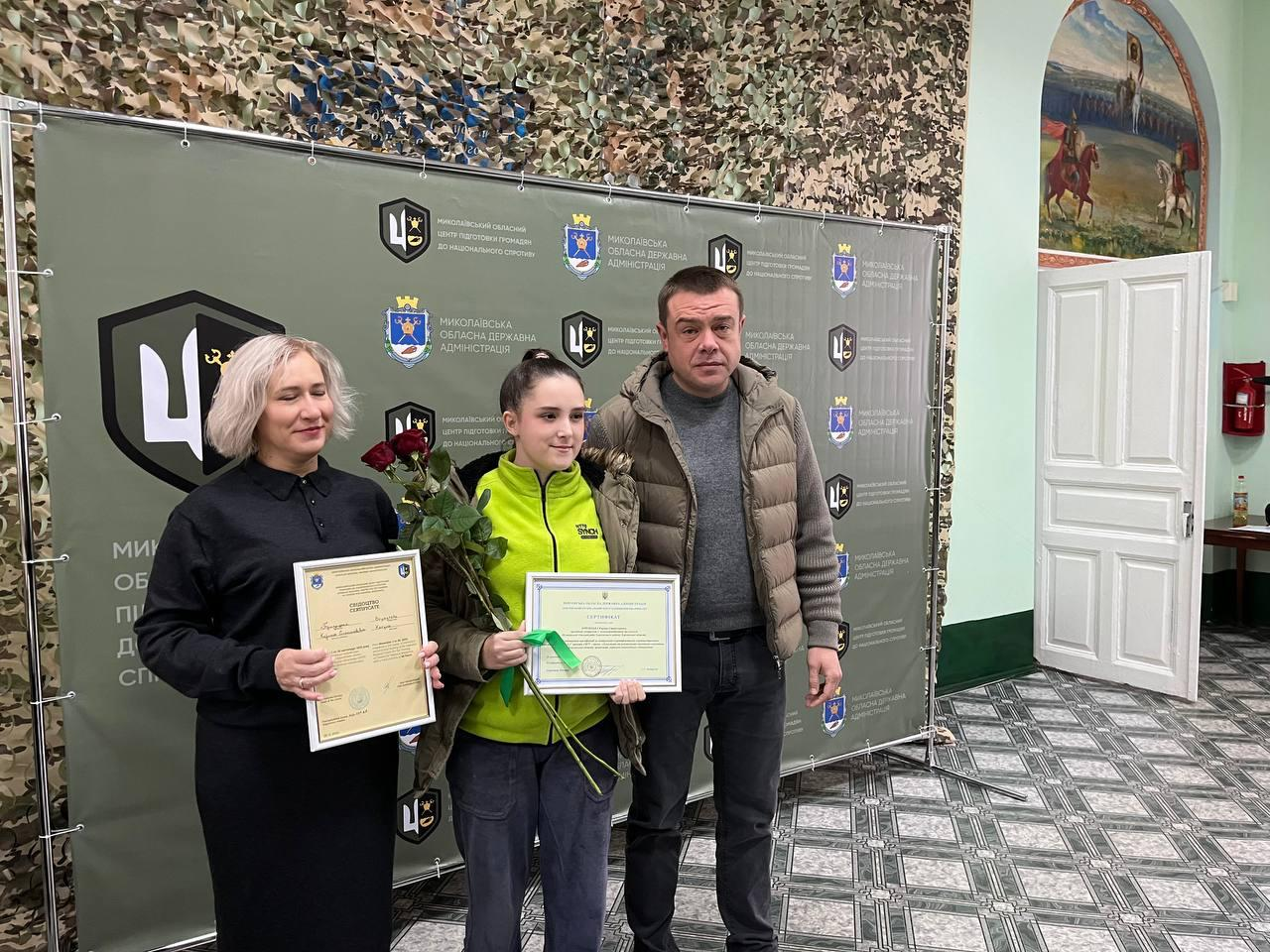 Груповий знімок жінки у чорному та чоловіка у жилетці з молодою дівчиною у спортивній куртці, яка тримає букет червоних троянд та документ. Вони стоять на тлі банера Миколаївської обласної державної адміністрації.