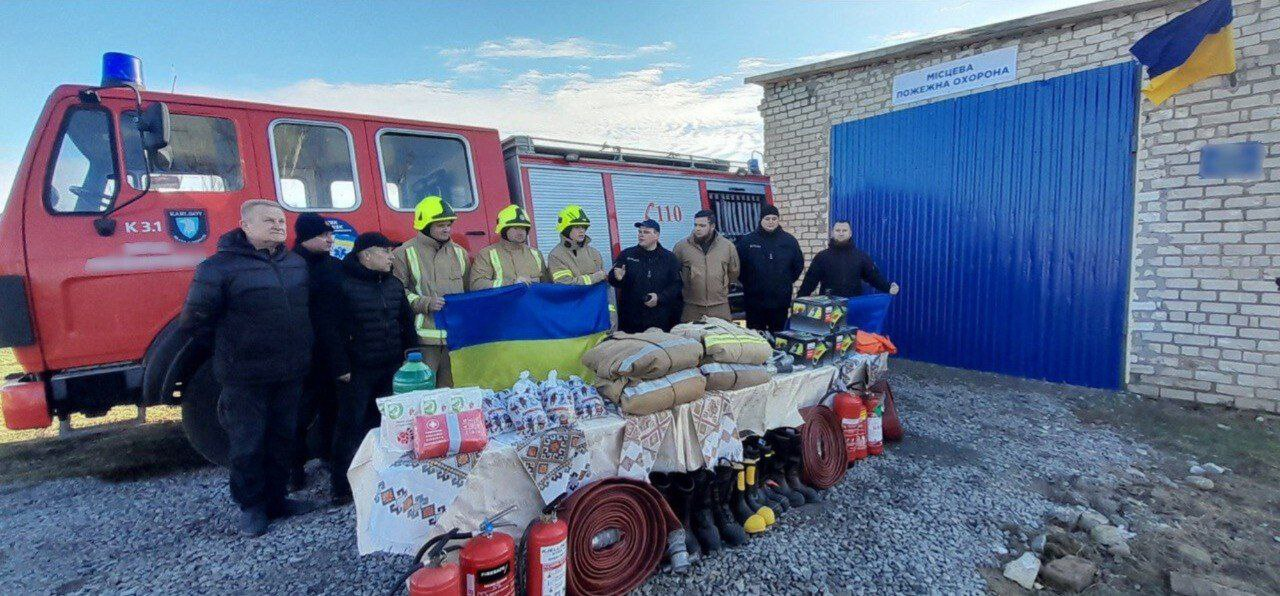 Група людей, які стоять перед червоною пожежною машиною та будівлею з синіми воротами, на яких видно напис: МІСЦЕВА ПОЖЕЖНА ОХОРОНА. Перед ними на столі викладена велика кількість пожежного та рятувального обладнання, включаючи пожежні рукави, вогнегасники, чоботи та захисні засоби. Кілька чоловіків у центрі тримають український прапор, а деякі одягнені в пожежні шоломи.